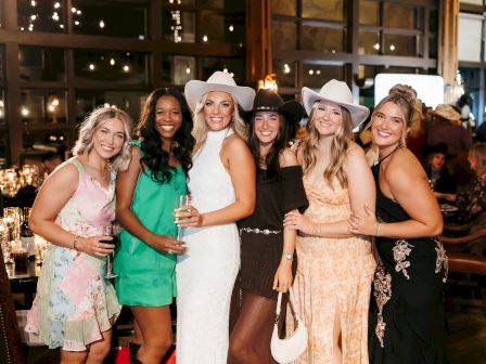 A group of six people smiling indoors, wearing dresses and cowboy hats, with warm lighting and a wooden interior setting.