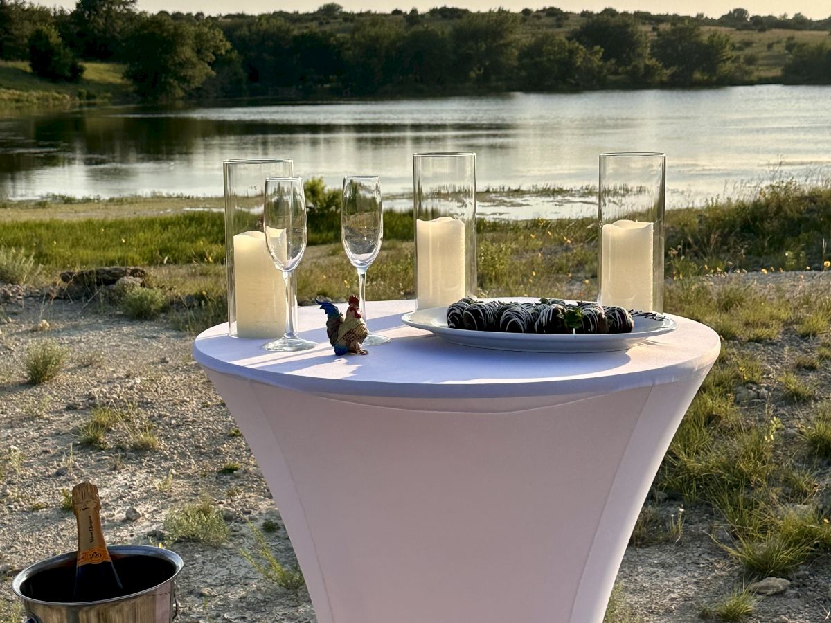 A cocktail table by a lake with drinks, glasses, and a bucket on a stand, set against a scenic outdoor backdrop.