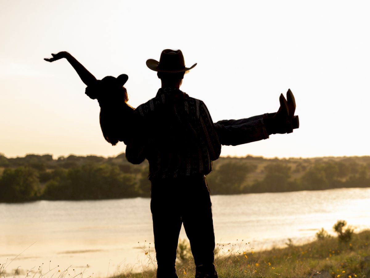 A silhouette of a person in a hat holding another person playfully by a riverbank, both against a bright sky.