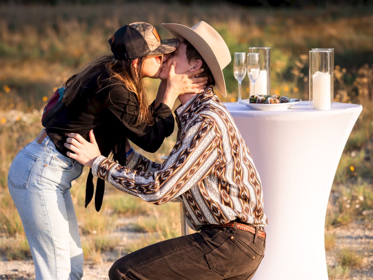 A couple is kissing outdoors, with the man kneeling. A table with glasses and a candle is nearby in a scenic setting.