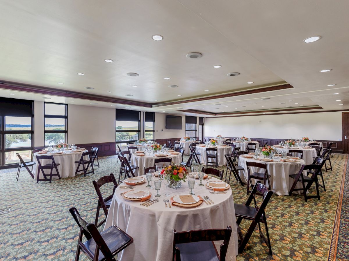 The image shows a banquet room with round tables set for an event, featuring white tablecloths, chairs, and centerpieces.