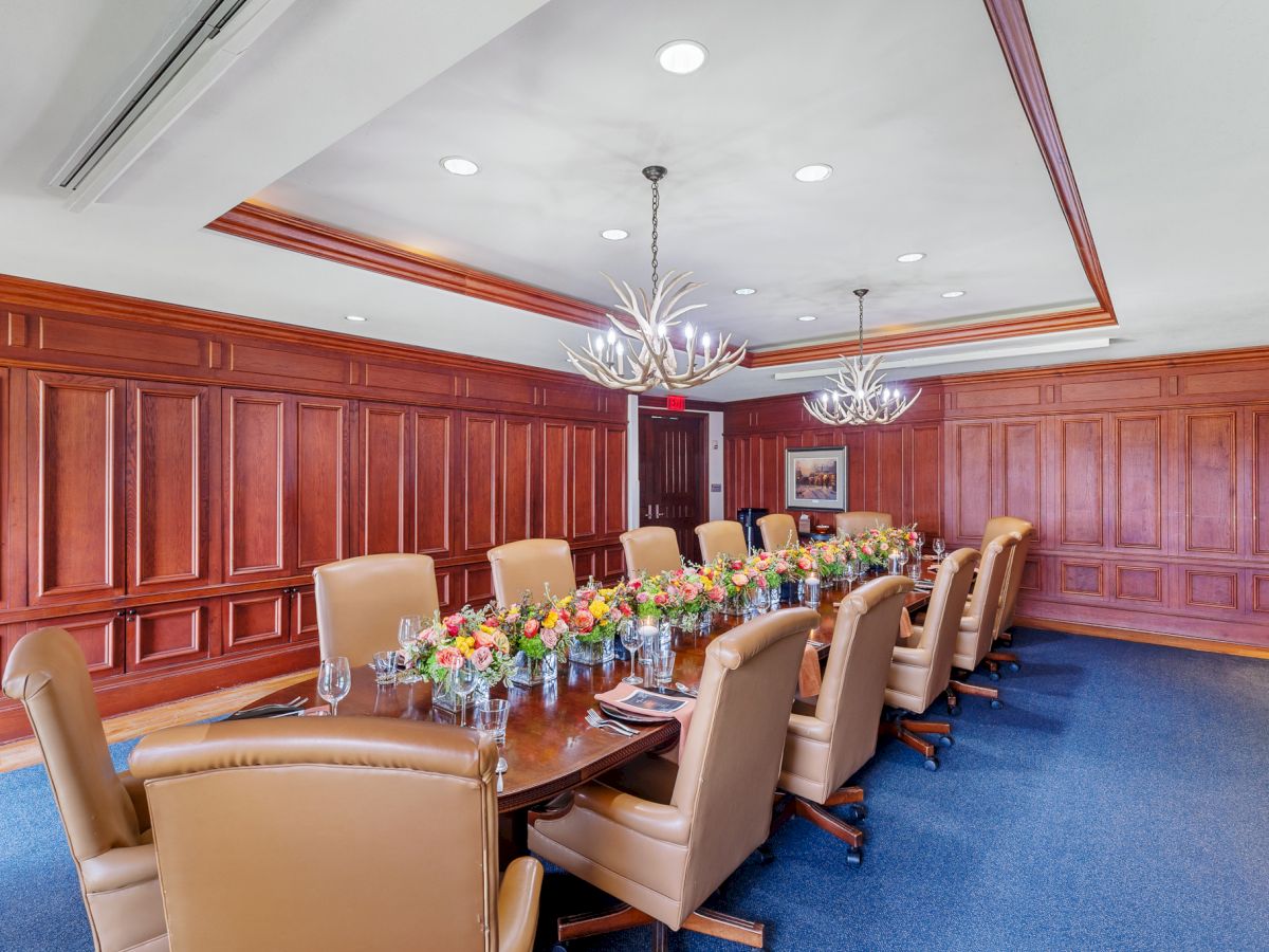 A formal conference room with a long table, leather chairs, floral centerpieces, wood paneling, and two chandeliers on the ceiling.