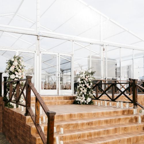 A clear tent with wooden stairs is adorned with floral arrangements and candle-lined steps, set outdoors for an event such as a wedding.