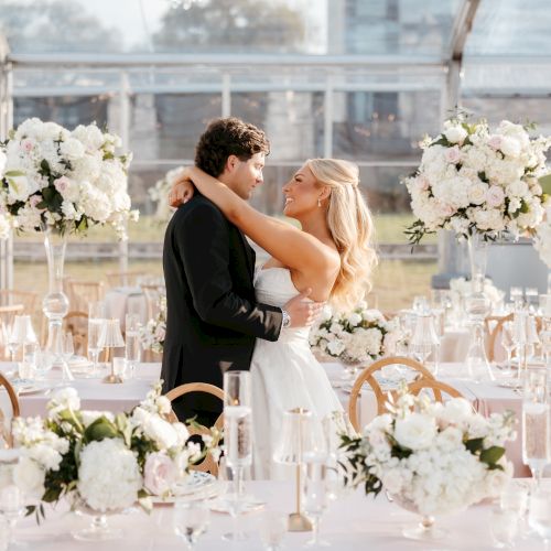 A couple embraces in a beautifully decorated, floral-filled wedding venue with elegant table settings and large white flower arrangements.