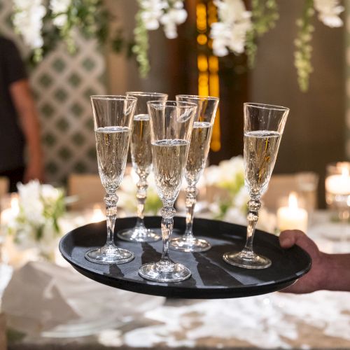 A tray with six champagne flutes filled with bubbly liquid is held by a person in a festive, elegantly decorated setting.