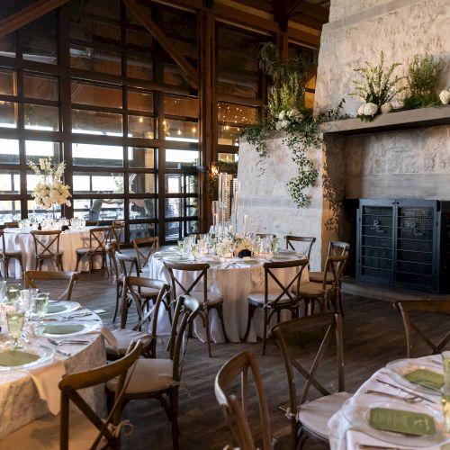 An elegantly decorated event space with round tables, wooden chairs, floral arrangements, and a fireplace, set for a gathering or celebration.