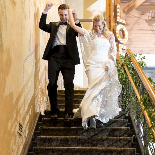 A couple in formal attire joyfully descends a staircase, hands raised in celebration, surrounded by warm lighting and floral decor.