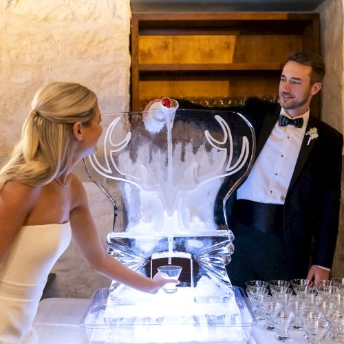 A person in formal attire pours a drink into a stag-shaped ice sculpture, while another person fills a glass below it.