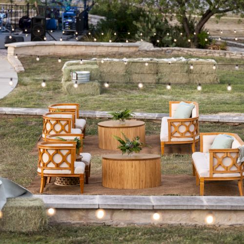 The image shows an outdoor lounge area with wooden chairs, round tables, and string lights, set on grass with a path and stage nearby.