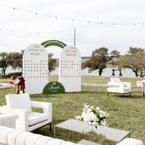 Elegant outdoor seating with white couches, a coffee table, and a decorative sign backdrop on a lawn by a body of water.