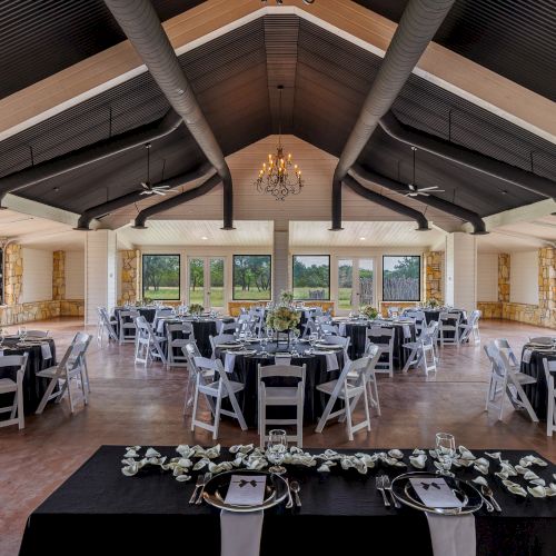 The image shows a decorated event venue with white chairs, round tables, and large windows, featuring a chandelier and black tablecloths.
