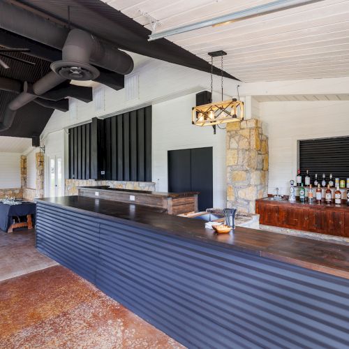 A spacious bar with a long counter, wooden accents, and various bottles displayed. Modern chandeliers hang from the ceiling.
