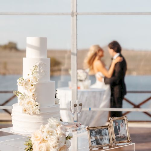 A white wedding cake with flowers, a framed photo, and a couple dancing in the background near a lake.