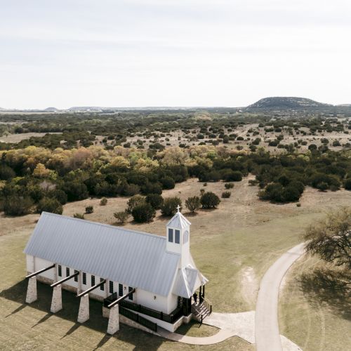 A white church with a steeple in a rural landscape, surrounded by greenery and hills under a clear sky.
