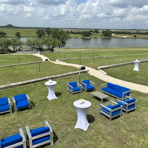 Outdoor seating area with blue chairs and white tables on grass, overlooking a pond and landscape under a cloudy sky.