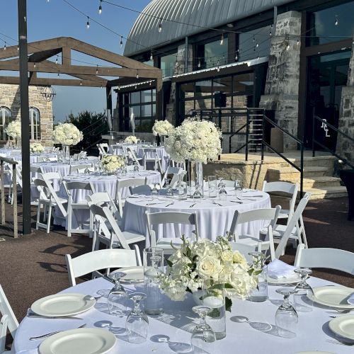 An outdoor event setup with white tables and chairs, floral centerpieces, and string lights near a modern building.