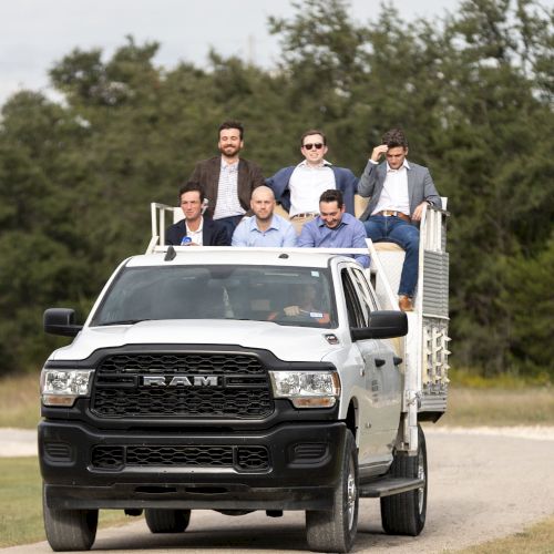 A group of people riding on top of a moving white Ram truck on a rural road.