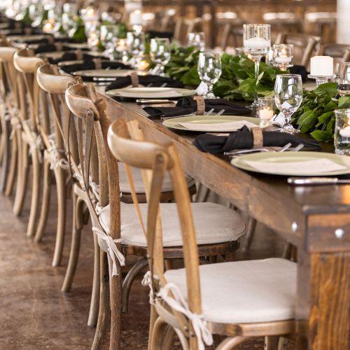 A long dining table set for an event, featuring wooden chairs, elegant tableware, and greenery centerpieces, awaits guests.
