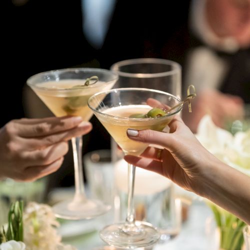 People are toasting with cocktail glasses at a formal event, surrounded by flowers and candles in a dimly lit setting.