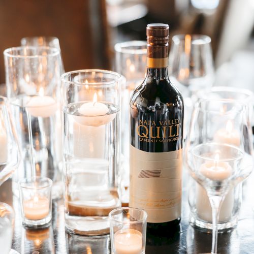 A wine bottle is surrounded by various glasses and lit candles on a table, creating a cozy ambiance.