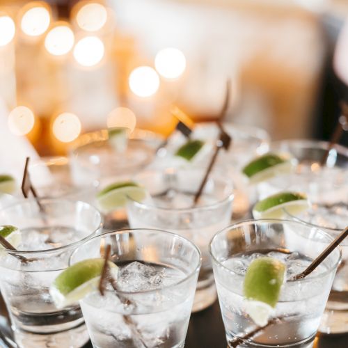 The image shows multiple glasses with clear liquid, lime wedges, and black stirrers on a tray, set against a bokeh light background.