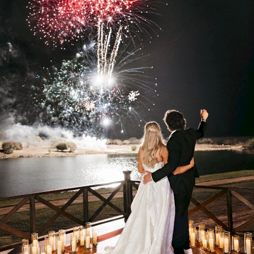 A couple watches a stunning fireworks display over a lake, surrounded by lit candles, on a wooden deck at night.