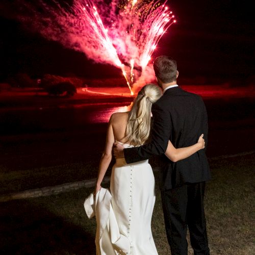 A couple dressed formally stands close together, watching vibrant fireworks illuminating the night sky.