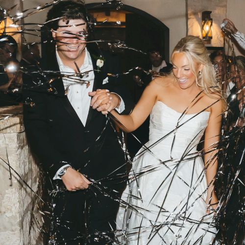 A bride and groom in formal attire are happily walking, surrounded by celebratory streamers, in a festive indoor setting.