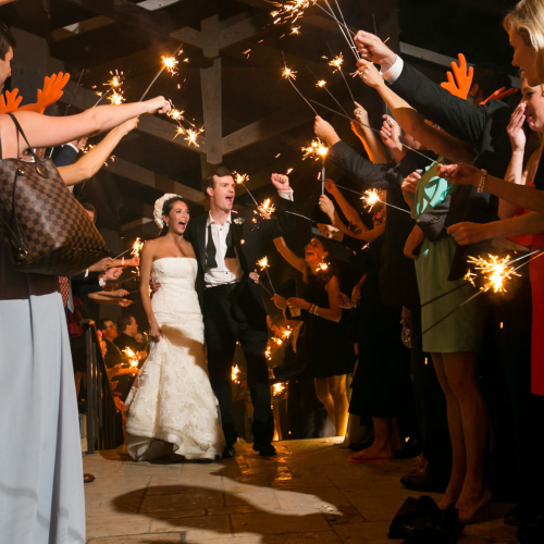 A bride and groom walk joyfully under an arch of sparklers held by guests, celebrating at a nighttime wedding event.
