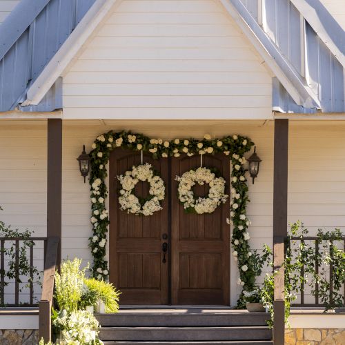 A building entrance with double wooden doors, adorned with white flower wreaths and garlands, steps, and plants on either side.