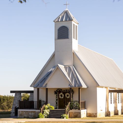 A small white chapel with a steeple, set in a sunny landscape with greenery. The building has a simple, rustic design.