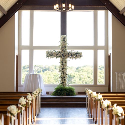 A beautifully decorated chapel interior with a large floral cross and pews adorned with white flowers, illuminated by sunlight.