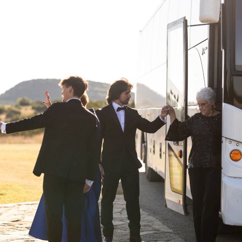 People in formal attire are near a bus, one is holding hands with an older woman. It appears to be a formal event or special occasion.