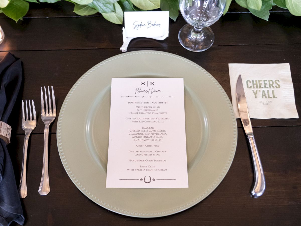 A table setting with a plate, menu, silverware, glass, and napkin. Green foliage decorates the table. A note reads "Cheers Y'all."