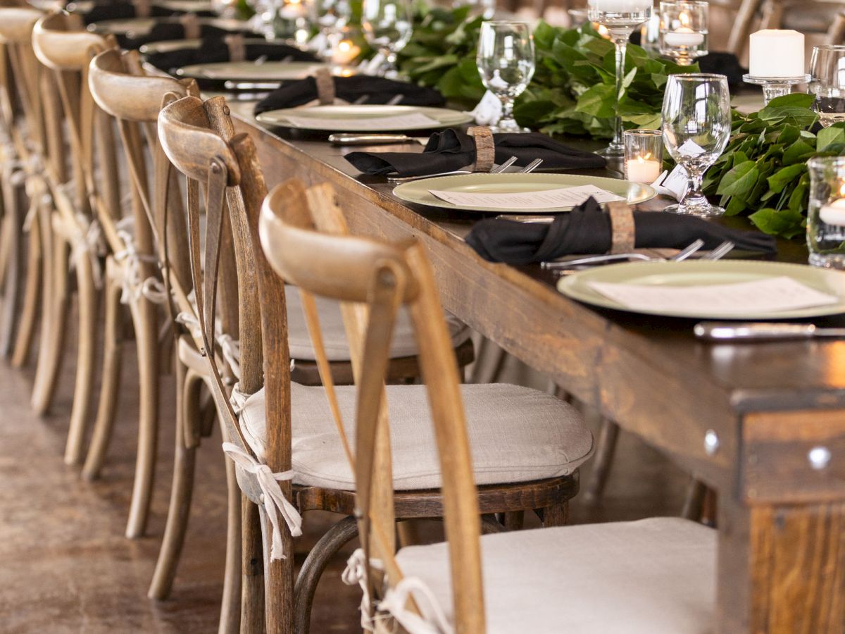 A long dining table set for an event with wooden chairs, black napkins, and greenery centerpieces, creating an elegant atmosphere.