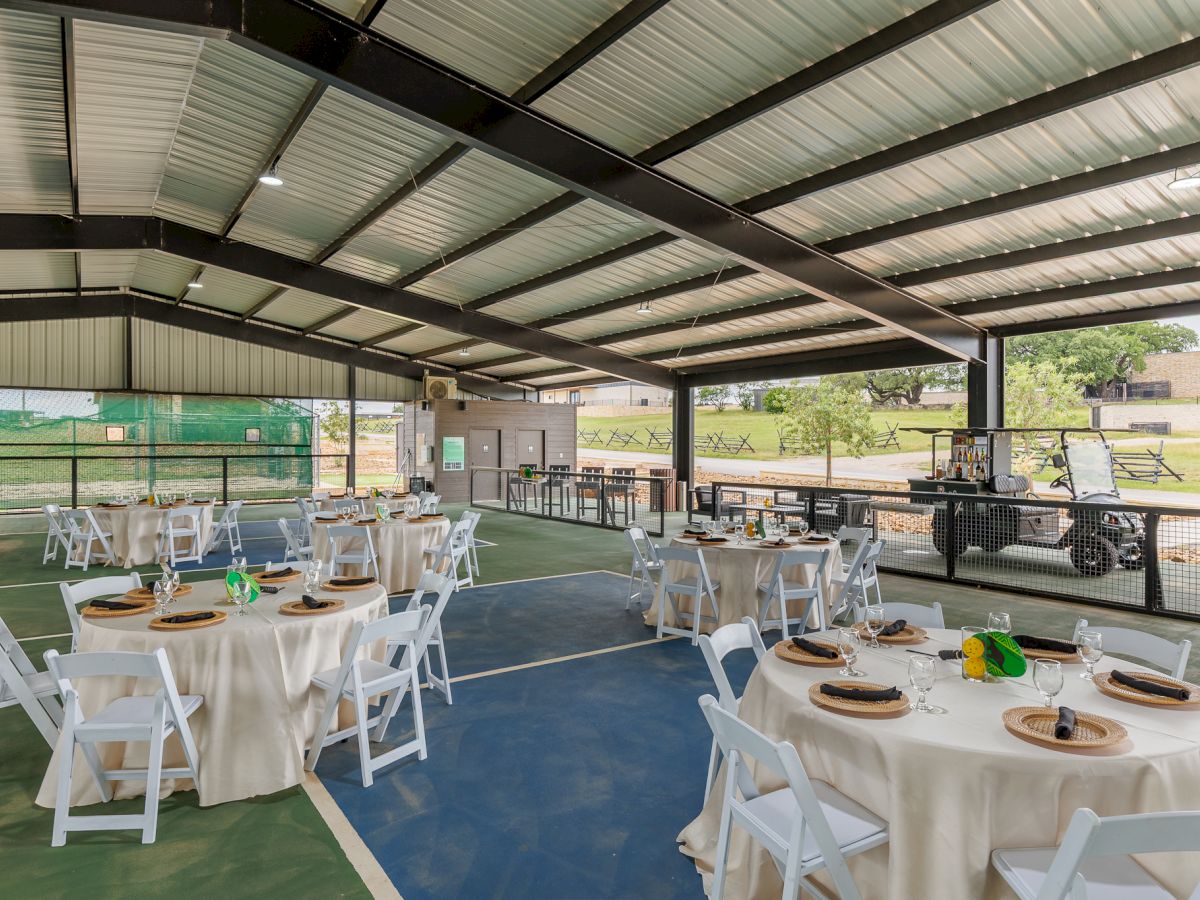 An outdoor venue with round tables, white chairs, and simple decorations under a metal roof, likely set up for an event or gathering.