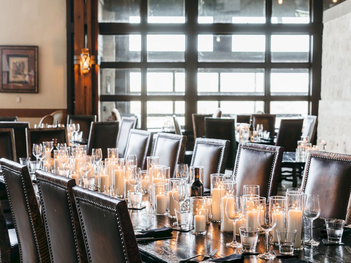 A long dining table set with numerous candles and glasses, surrounded by dark chairs in a bright room with large windows.