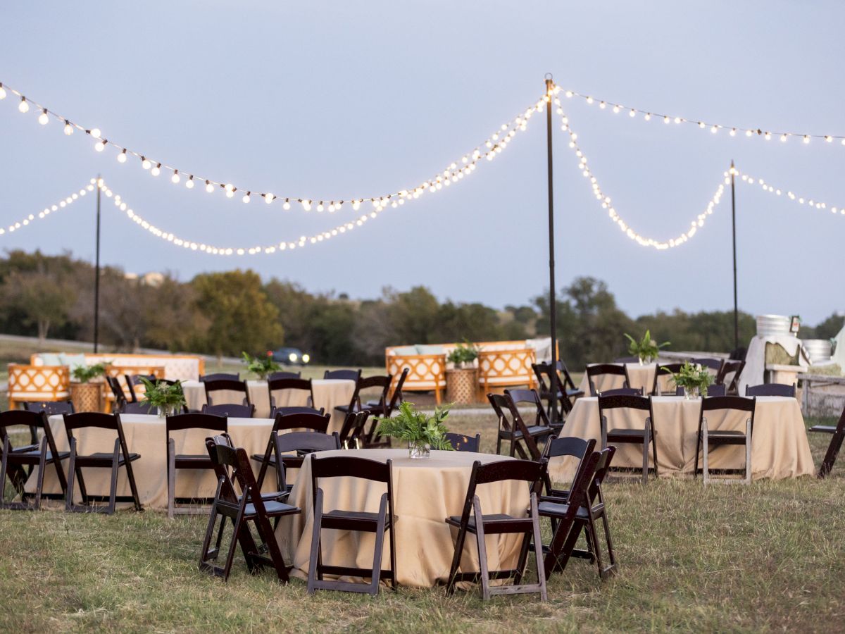 An outdoor event setup with round tables and chairs, string lights overhead, and a natural background at dusk.