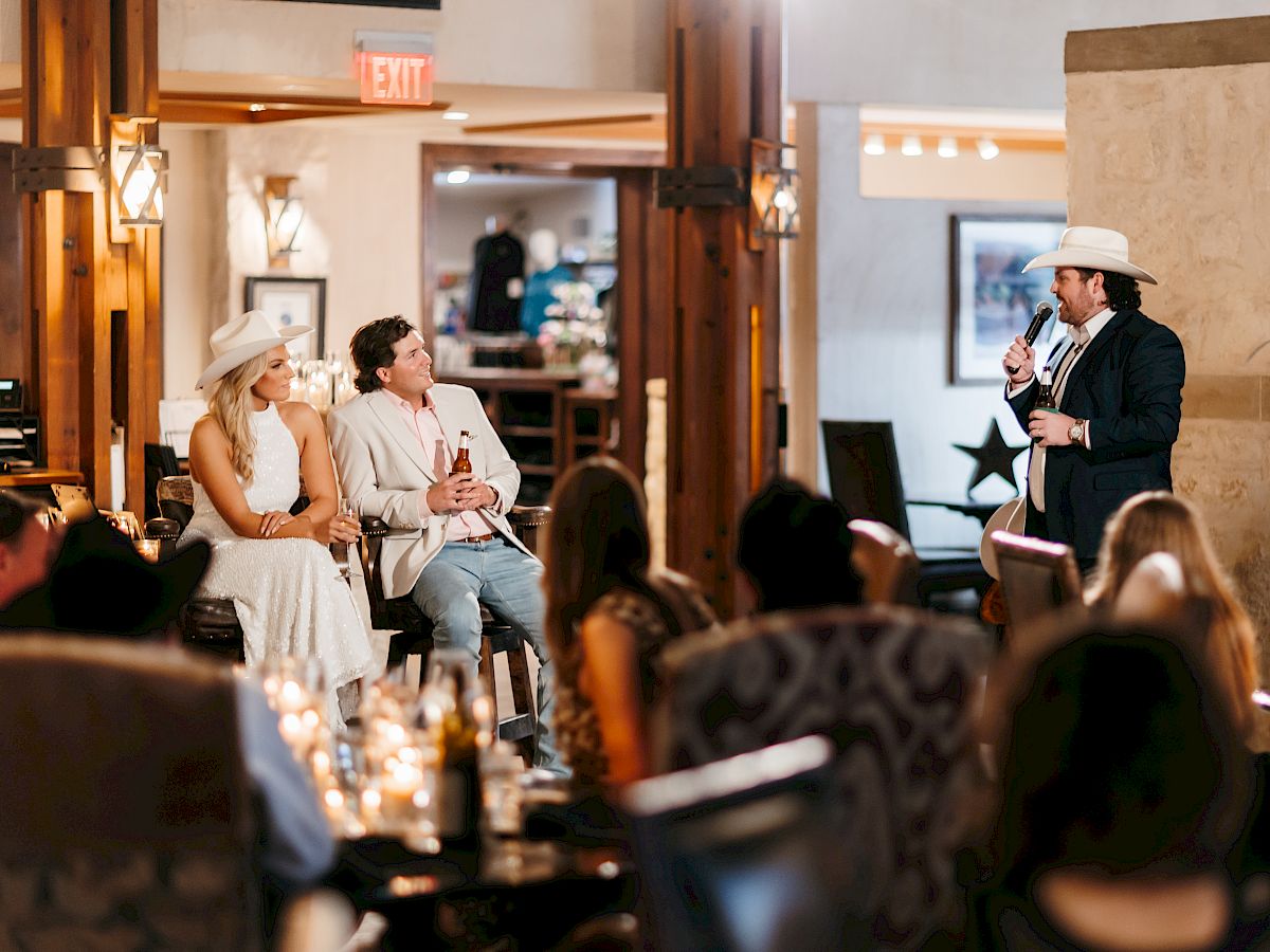 A person in a suit and cowboy hat speaks to a seated couple, also in hats, in a warmly lit indoor setting with an audience.