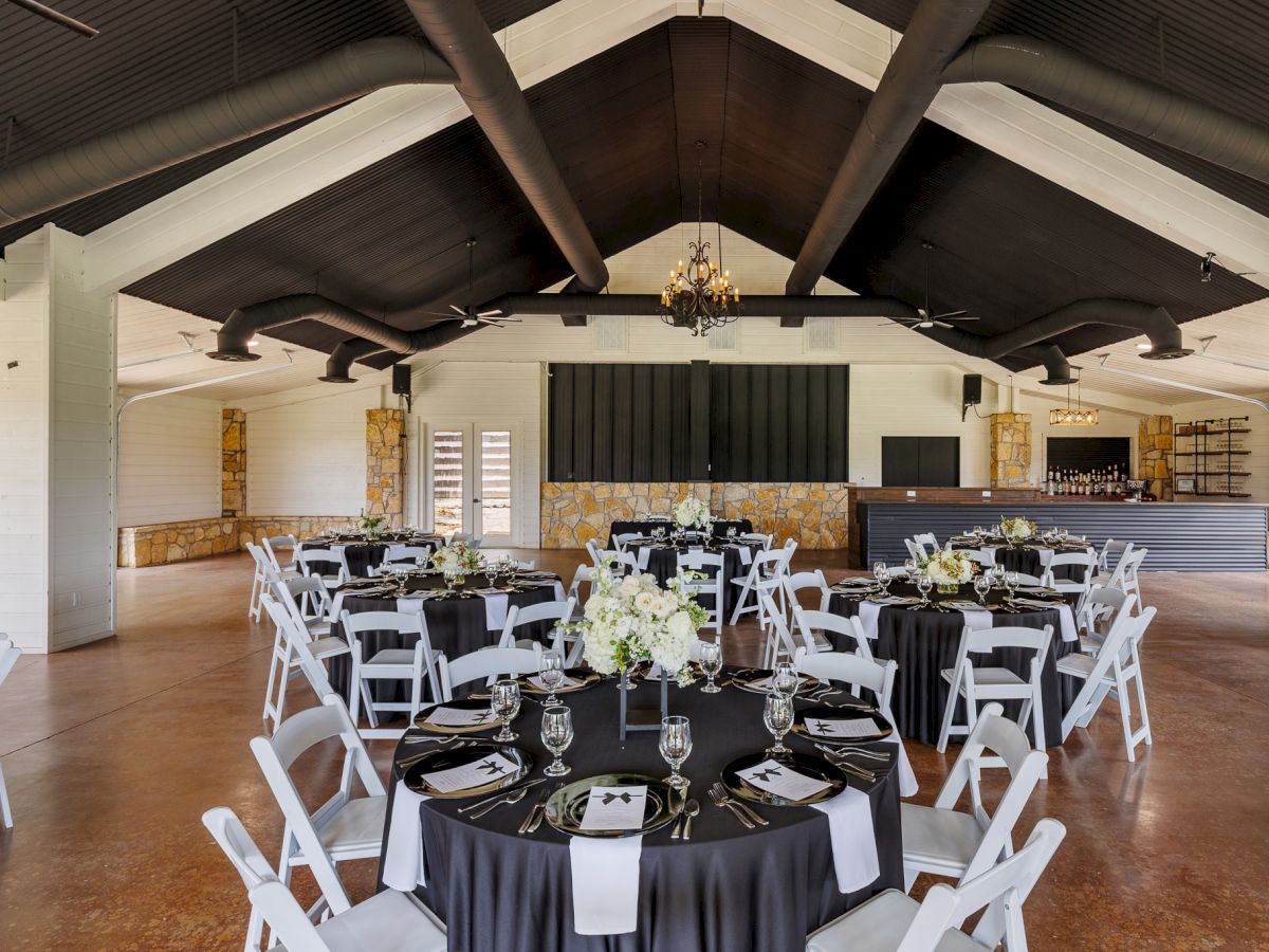 The image shows a decorated event hall with round tables set for dining and chairs arranged around them, beneath a high ceiling.