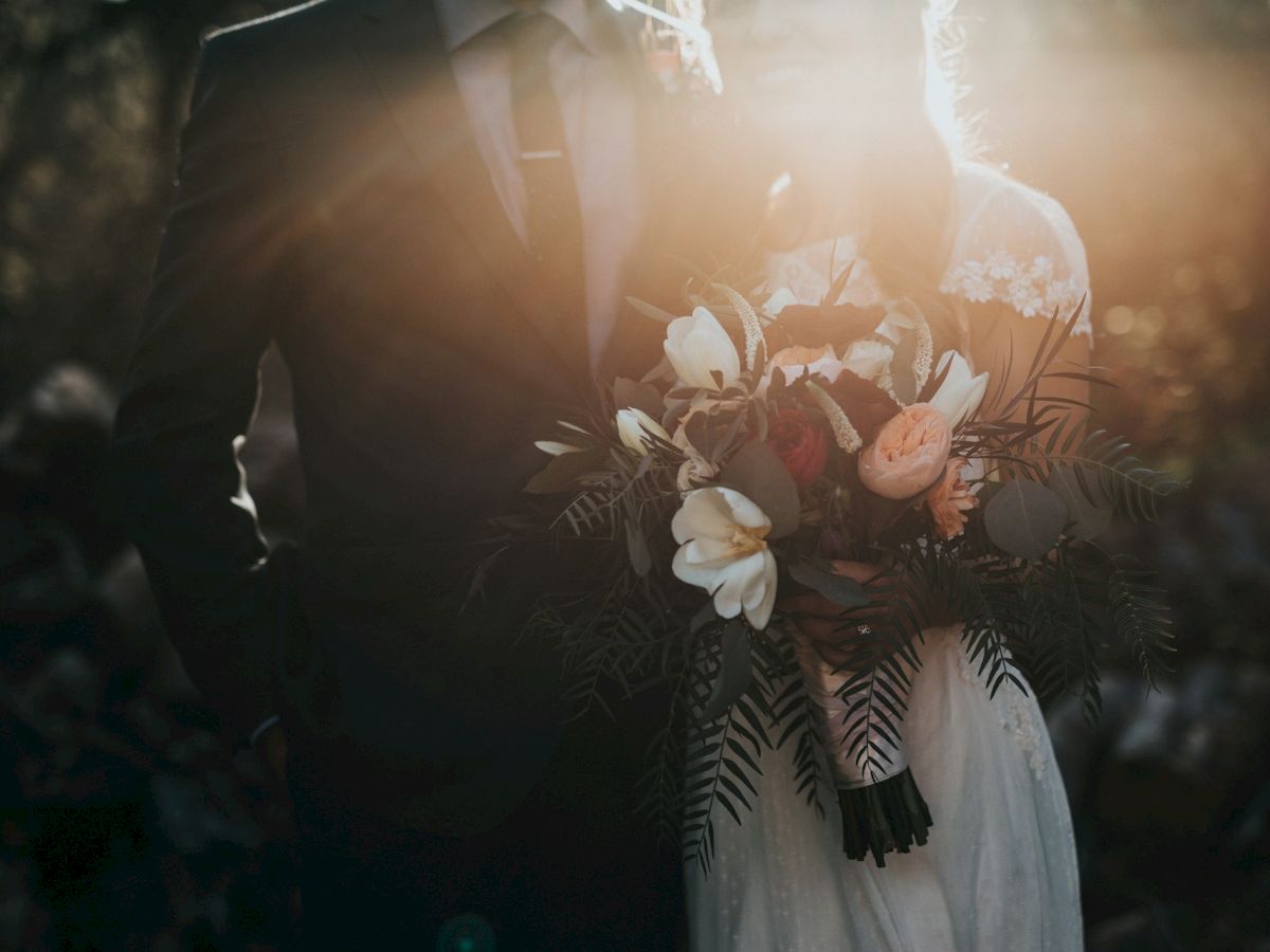 A couple stands closely, with the sun illuminating them, holding a bouquet of flowers.