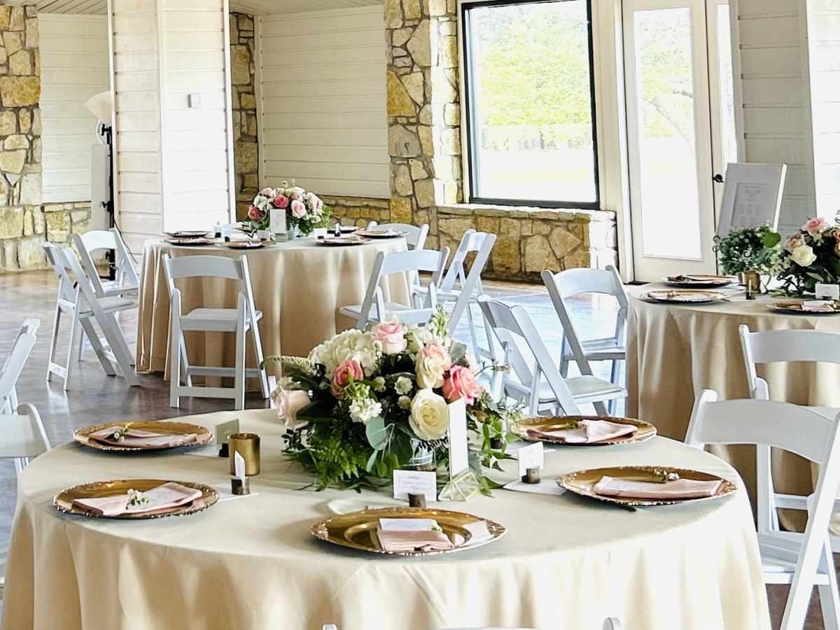 Elegant tables with beige tablecloths, floral centerpieces, and white chairs are set up for an event in a bright, spacious room.