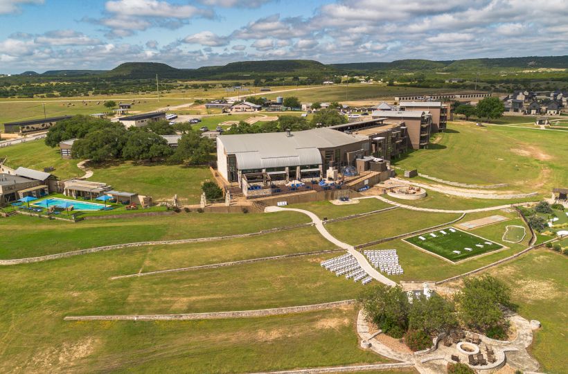 The image shows a sprawling landscape with a large building complex, green fields, pathways, a pool, and multiple outdoor seating areas.