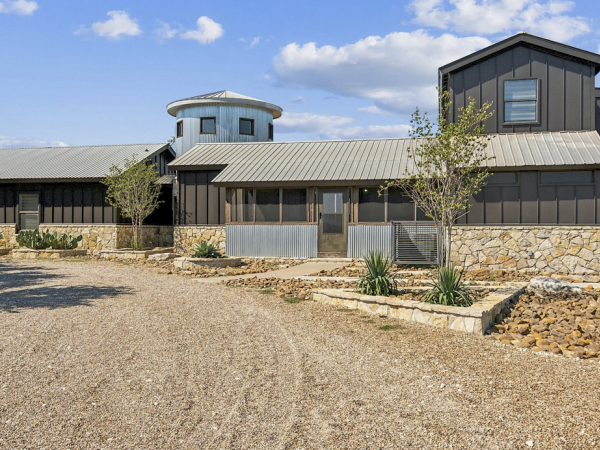 A modern barn-style building with metal and stone elements, large windows, surrounded by small trees and a gravel driveway.