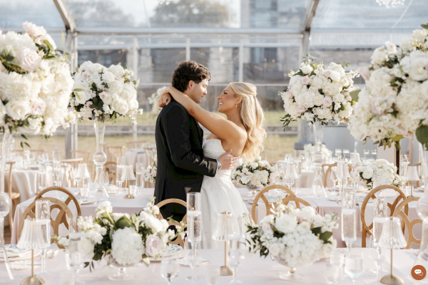 A couple embraces at an elegant wedding reception with white floral centerpieces and a transparent tent setting.