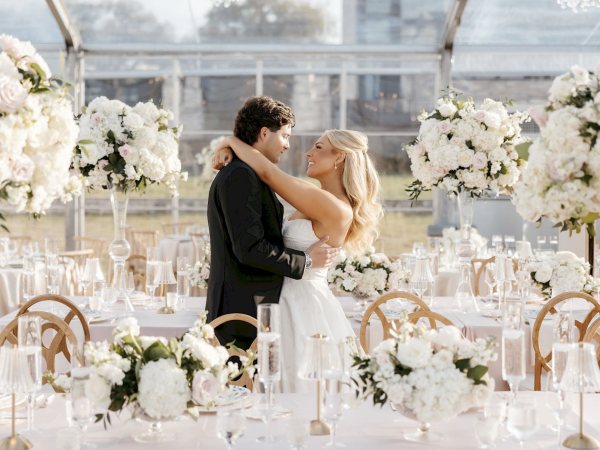 A couple embraces at an elegant wedding reception with white floral centerpieces and a transparent tent setting.