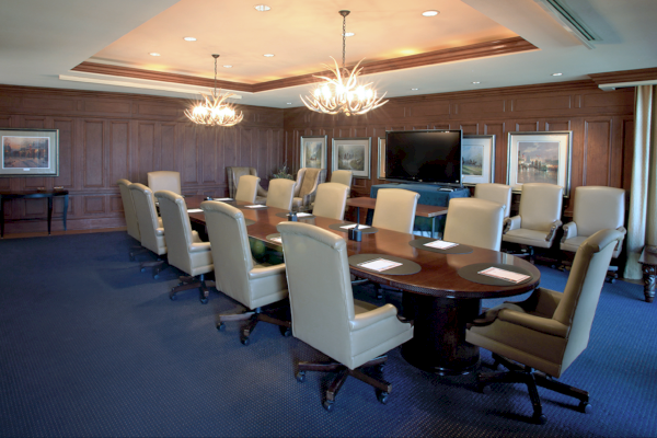 A boardroom with a long table, chairs, chandeliers, artwork, and a large screen is visible.