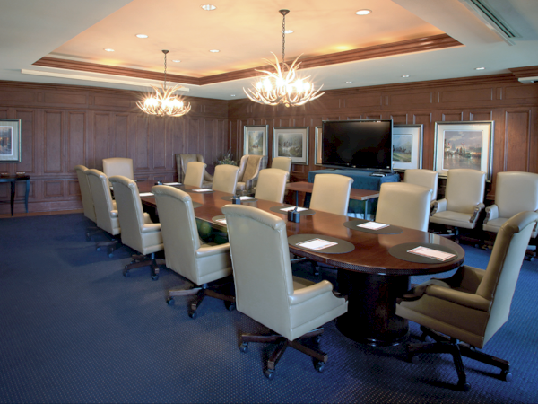 A boardroom with a long table, chairs, chandeliers, artwork, and a large screen is visible.