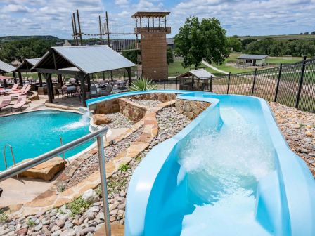 The image shows a blue waterslide leading into a pool, surrounded by a fence and patio area with lounge chairs, against a rural backdrop.