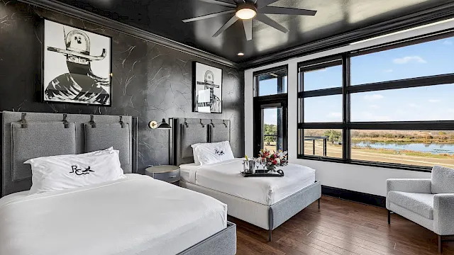 Modern hotel room with two beds, wall art, ceiling fan lights, and big windows showing a river view.
